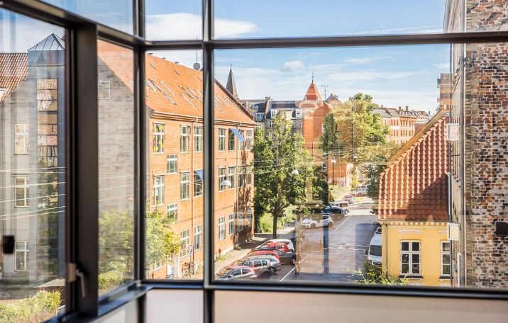 Kontor med udsigt til gaden og parkerede biler på Nørrebro. Bygningen med røde tag og flere vindueruder har skov i baggrunden og er beliggende på en højde
