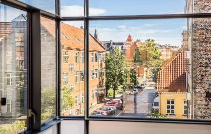 Kontor med udsigt til gaden og parkerede biler på Nørrebro. Bygningen med røde tag og flere vindueruder har skov i baggrunden og er beliggende på en højde