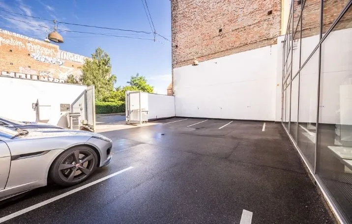 En silver Jaguar F-Type parkeret i en parkeringsplads med en bygning med glasvinduer og en murstensbygning bagved