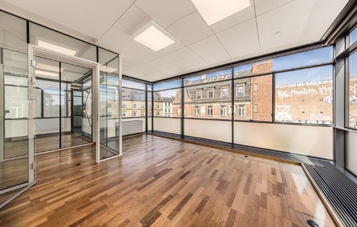 Glasvinduer og gulv i træ med et lyseloft i loftet og udsigt til bygninger udenfor