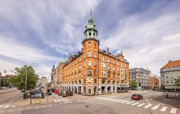 Klassisk og moderne bygning i København med hjørnemødelokale og åbent kontormiljø. Stort kontor med 258,40 m², 3 kontorer og plads til ca. 24 medarbejdere. Parkerede biler og grønne træer.