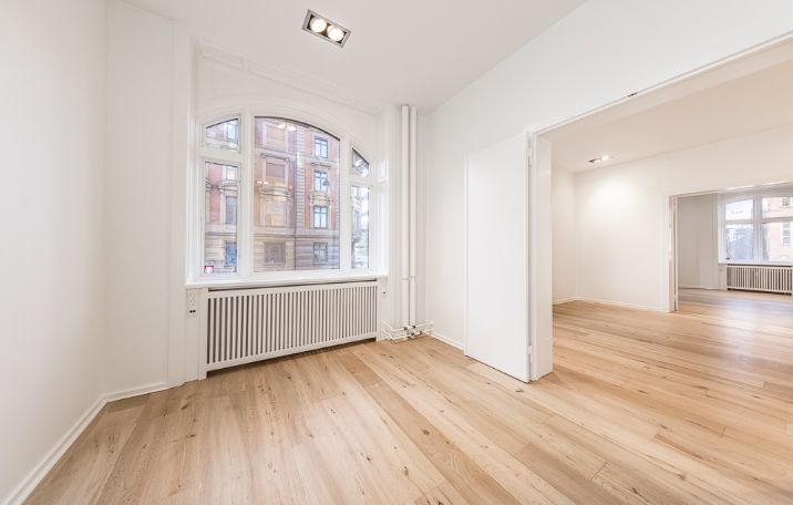 Moderne kontor med hjørnemødelokale og lyse gulve. Vinduer med udsigt til bygning med mange vinduer. Radiator og lysekroner.