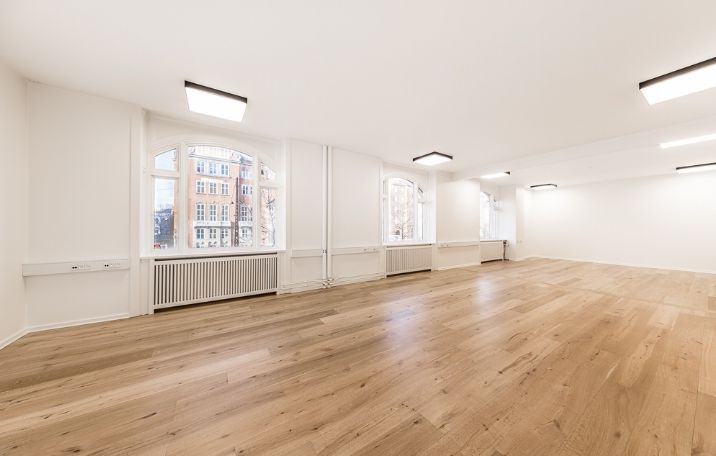 Lys og rummelig kontor med lysegule gulvplanker og hvide vægge. Store vinduer med masser af dagslys. Moderne møbler og radiatorer under vinduerne