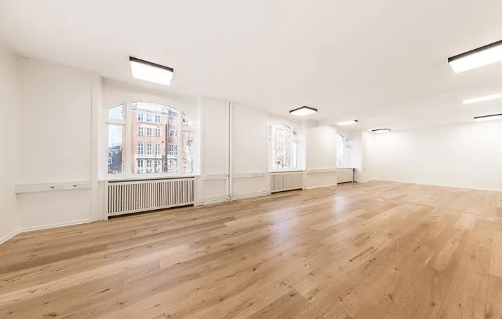 Lys og rummelig kontor med lysegule gulvplanker og hvide vægge. Store vinduer med masser af dagslys. Moderne møbler og radiatorer under vinduerne