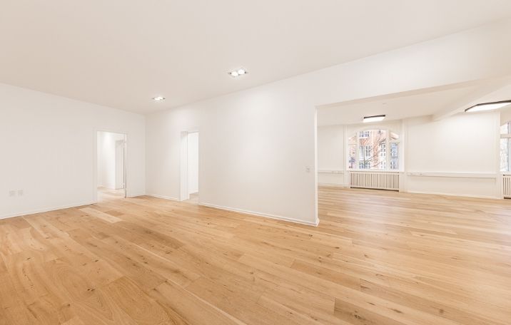 Et klassisk kontor med hvide vægge og lysegult trægulv. Plads til flere medarbejdere og lyse loftslamper. Et hjørnemødelokale med vinduer og radiatorer.