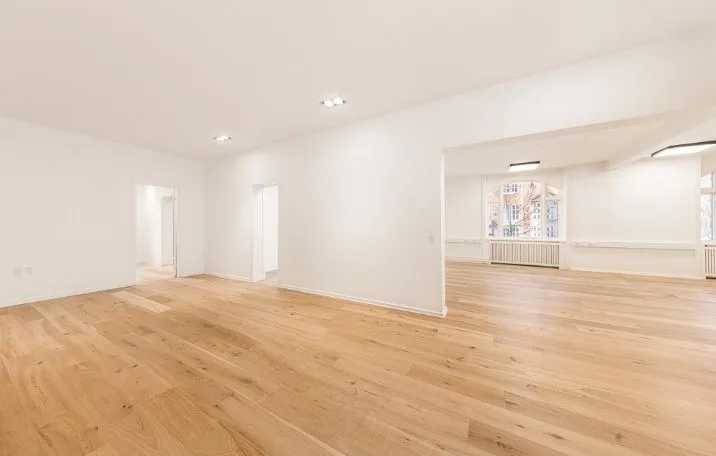 Et klassisk kontor med hvide vægge og lysegult trægulv. Plads til flere medarbejdere og lyse loftslamper. Et hjørnemødelokale med vinduer og radiatorer.