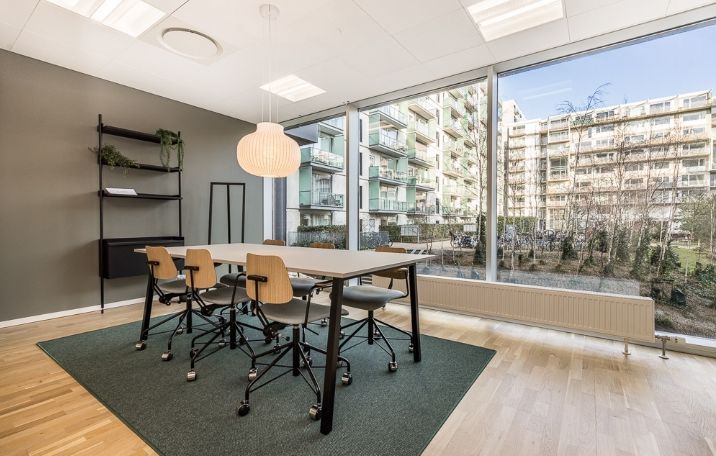 Kontor til leje med lyse møbler og høj vinduesfront mod grønne træer i baggrunden på Arne Jacobsens Allé 7, 1., 2300 København S