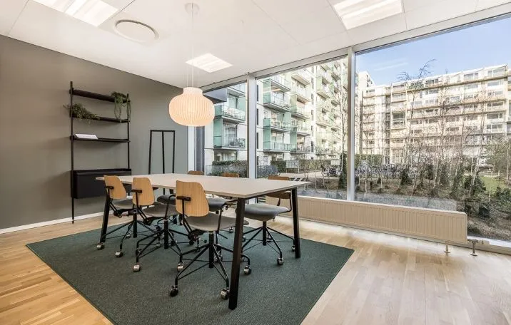 Kontor til leje med lyse møbler og høj vinduesfront mod grønne træer i baggrunden på Arne Jacobsens Allé 7, 1., 2300 København S