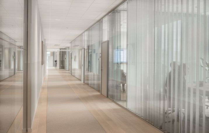 Langt, tomt kontorhjørne med glas og hvidt tapet, højt gulv, moderne indretning, stærkt lys, personer sidder ved borde, mødelokale, søjler, gulvbelægning, vinduer, kontorlokaler