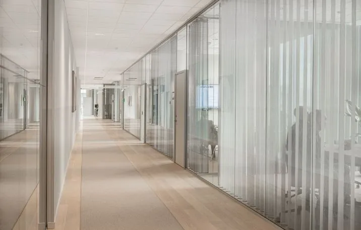 Langt, tomt kontorhjørne med glas og hvidt tapet, højt gulv, moderne indretning, stærkt lys, personer sidder ved borde, mødelokale, søjler, gulvbelægning, vinduer, kontorlokaler