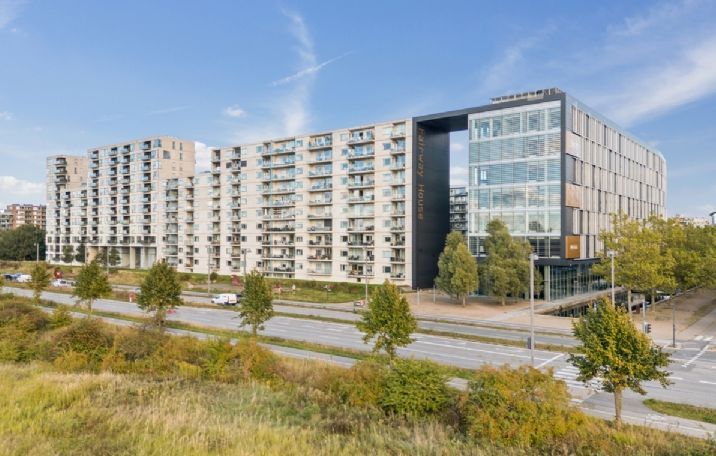 Moderne kontorbygning med glasfacade på Arne Jacobsens Allé, omgivet af grønne træer og buske, med bilparkeringsplads og brede veje