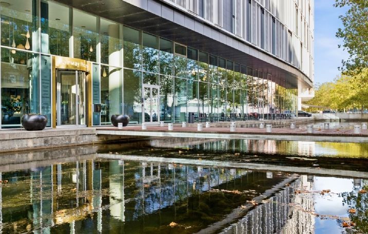En moderne bygning med glasfassade og sø foran, samt store, sorte boliger. Et stort gult skilt med teksten "Airway Hub"
