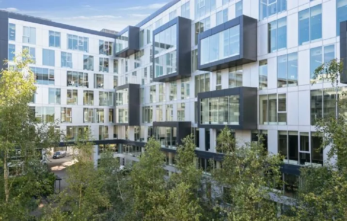 Kontorbygning med glasvinduer og metalkældre på Arne Jacobsens Allé 7 i København. Moderne design med grønne træer og et parkerede bil foran.