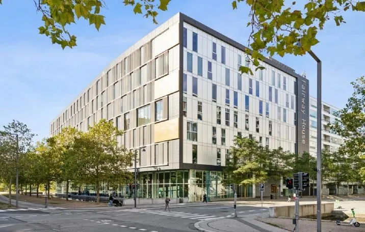 Moderne kontorbygning med store glasvinduer på Arne Jacobsens Allé. Træer og gågade. Personer går i baggrunden. Moderne trafiklys. Bygningen er lyse og rummelige.