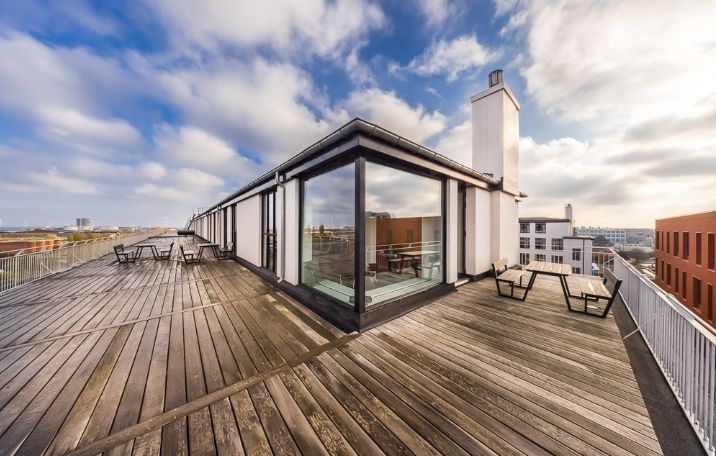 Terrasse på 5. sal med udsigt over København. Værelse med trægulv og glasvægge. Bord og stole udenfor. Lys og solrige himmel med skyer.