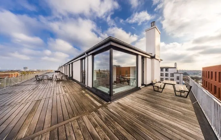 Terrasse på 5. sal med udsigt over København. Værelse med trægulv og glasvægge. Bord og stole udenfor. Lys og solrige himmel med skyer.