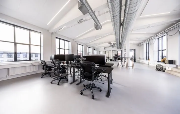 Moderne kontor med høje ruder og lyse lofter, fuld af ergonomiske kontorstolar og borde med computere og skrivebordslamper