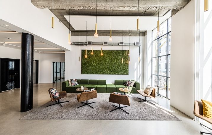 Kontor med grøn sofa og stole i lobbyen. Lamper hænger fra loftet. Vindue med udsigt til byen. Plantevæg og højloftede rum med lyse loftsbjælker.