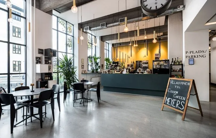 Kontor med moderne interiør og udsigt over byen. Vægge med hylder og lysekroner. Et bord med et tavle og en tekst på det. En plante og en sofa med en pude på gulvet.