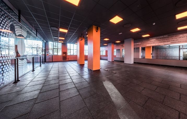 Fitnesscenter med moderne interiørdesign, højt loft, søjler og gulvbelægning, der reflekterer lys fra lysekroner. Væggen er dekoreret med skrift og mønstre