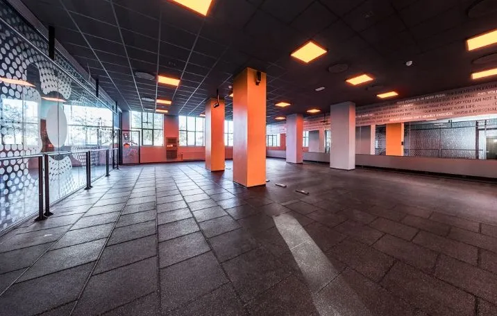 Fitnesscenter med moderne interiørdesign, højt loft, søjler og gulvbelægning, der reflekterer lys fra lysekroner. Væggen er dekoreret med skrift og mønstre