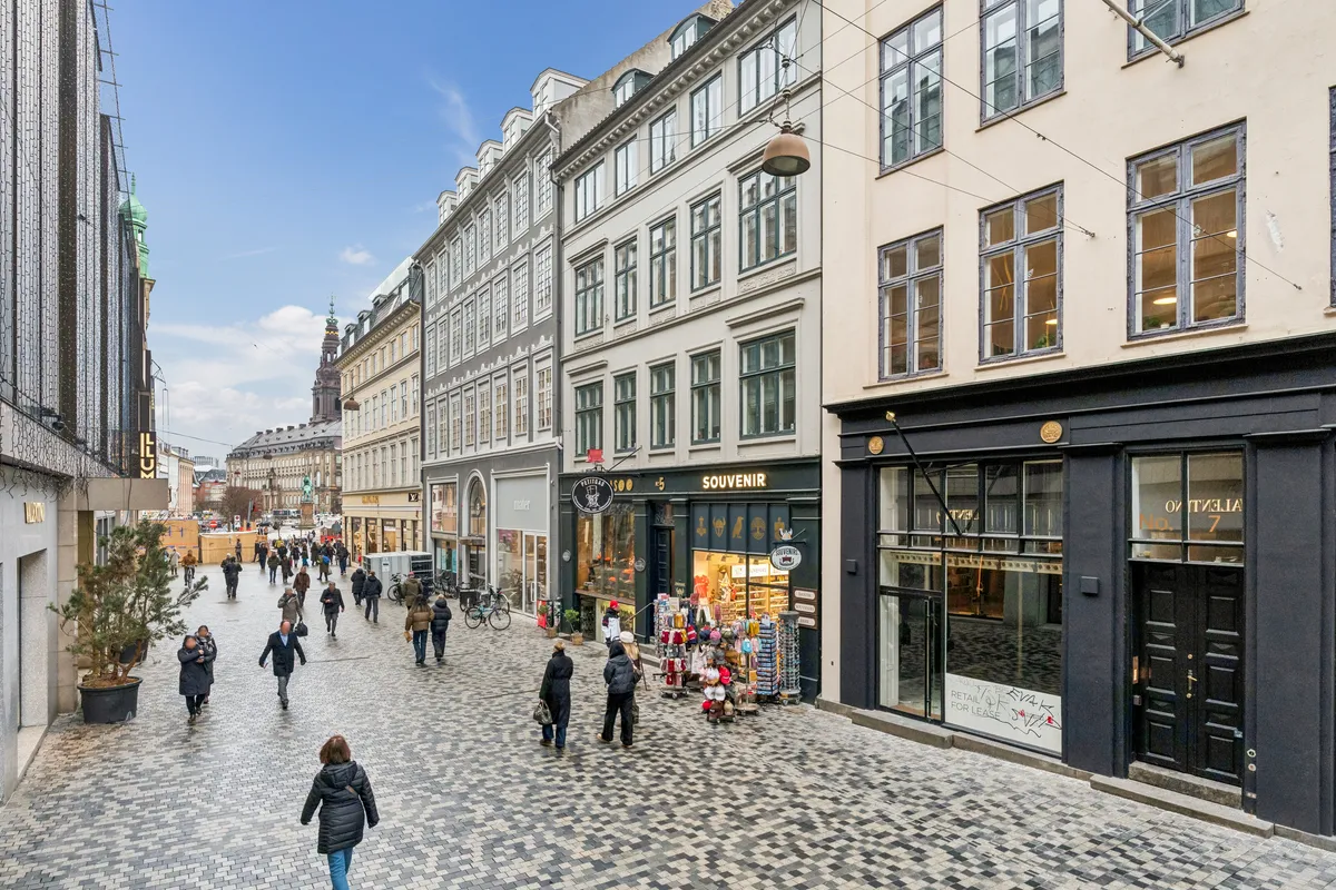 Mennesker vandrer på en middelalderlig brostensgade med forretninger og en søjle med en sognekirke i baggrunden, alle på Købmagergade i København
