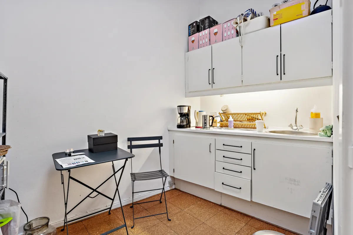 Kontor til leje på Købmagergade 3B, 4., 1150 København K. Slank bord med hængestolle. Højre er køkken med apparater og træbord. Loftsskabe med flasker og kasser.