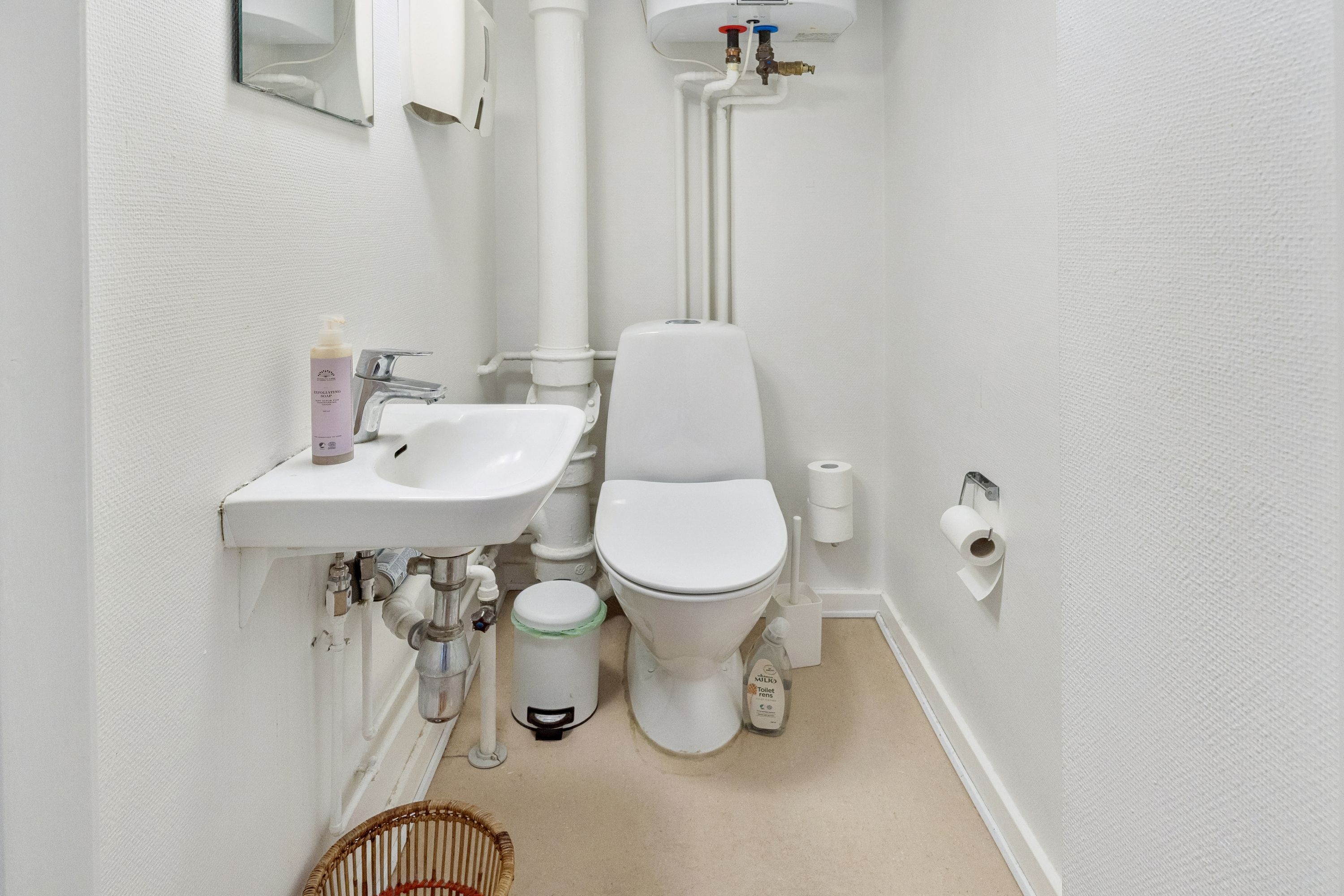 En lille hvid toiletrum med en hvid toilet, et hvidt vask og en hvid toiletpapirholder. Et toiletpapir er hængende på holderen. Et støjpose og en håndvask med sæbe er på bordet.