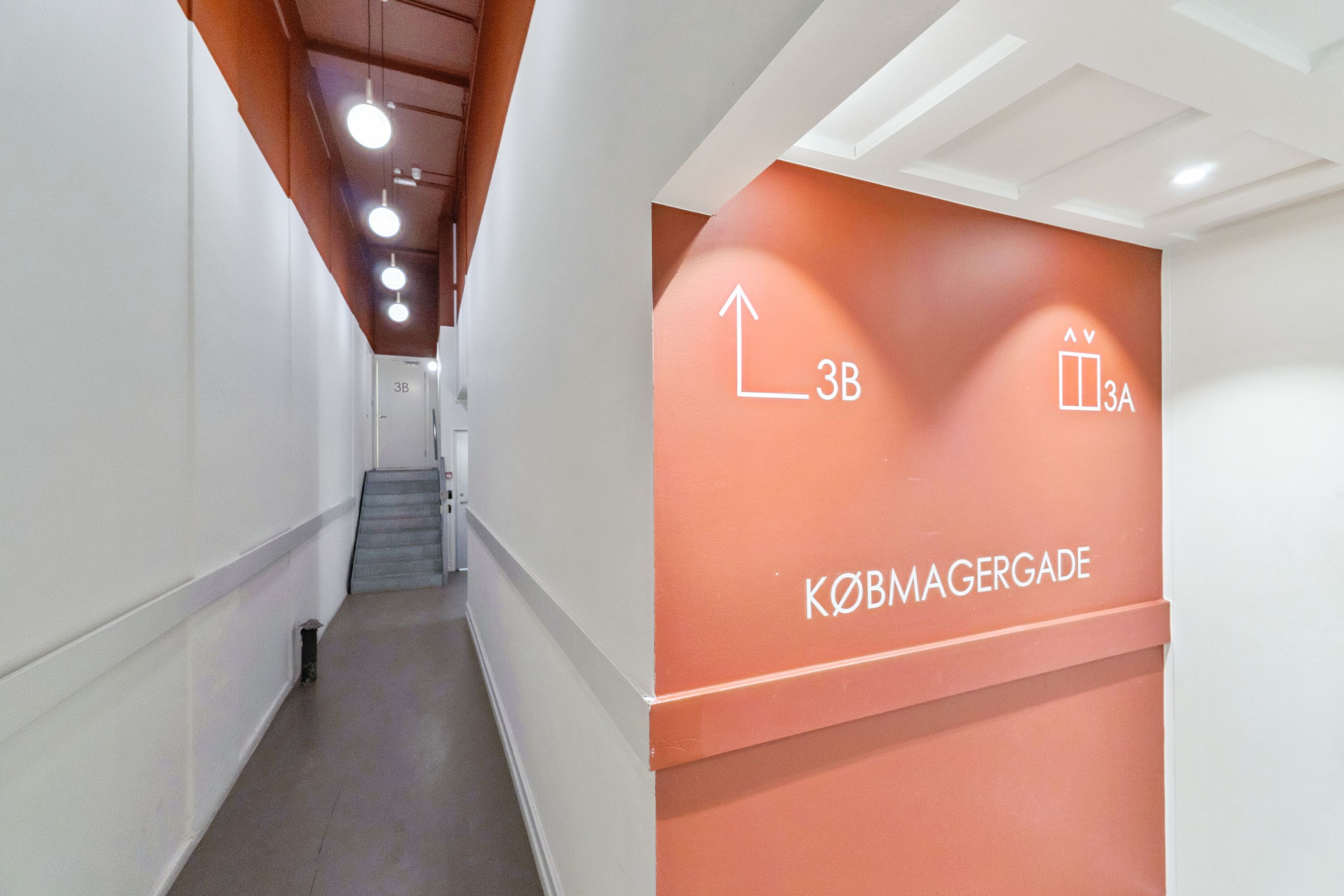 Kontor til leje på Købmagergade 3B i København. Gangen er møbleret med hvide vægge og lysekroner. Der er en hvid dør og trapper i baggrunden.
