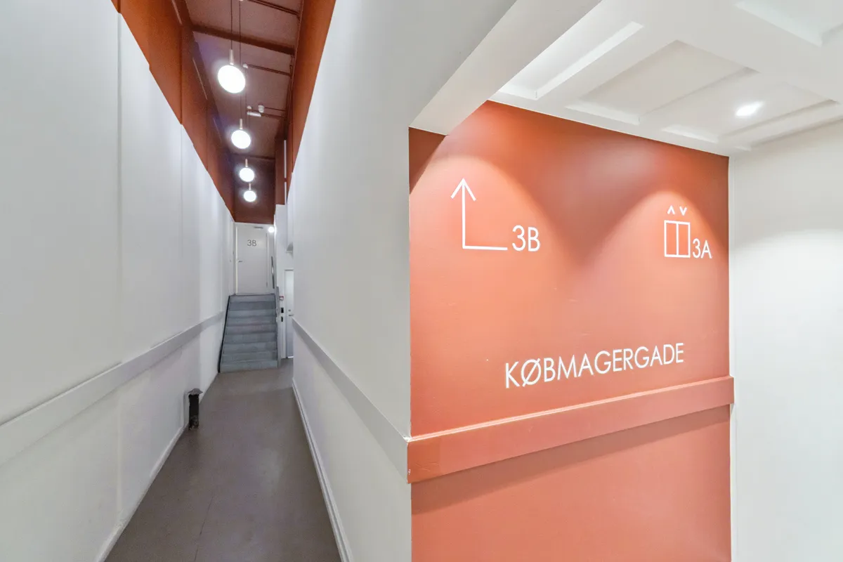 Kontor til leje på Købmagergade 3B i København. Gangen er møbleret med hvide vægge og lysekroner. Der er en hvid dør og trapper i baggrunden.