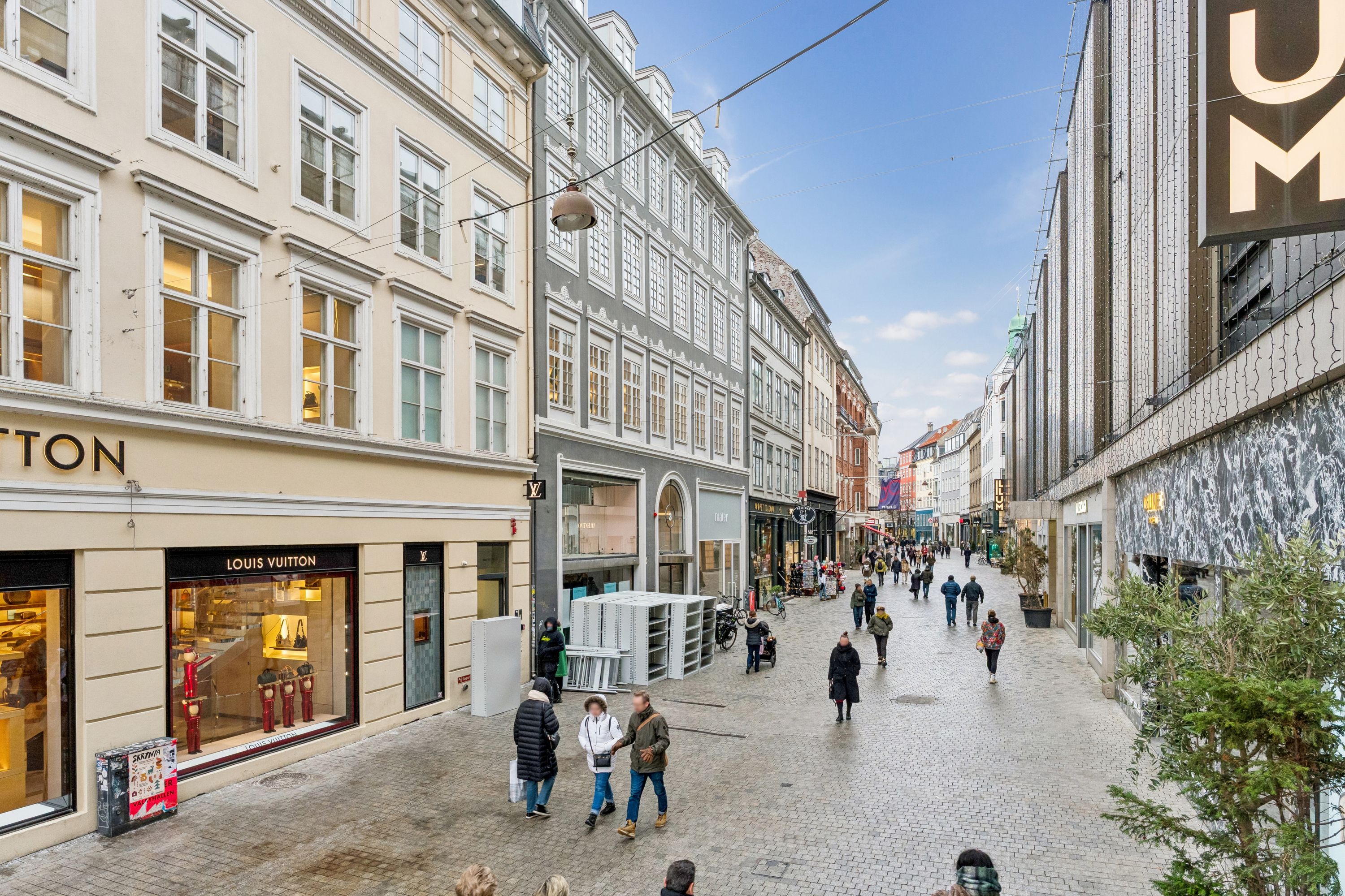 På Købmagergade går gående folk forbi butikker og Louis Vuitton butik og andre bygninger med lyse vinduer og en grøn plante på gaden