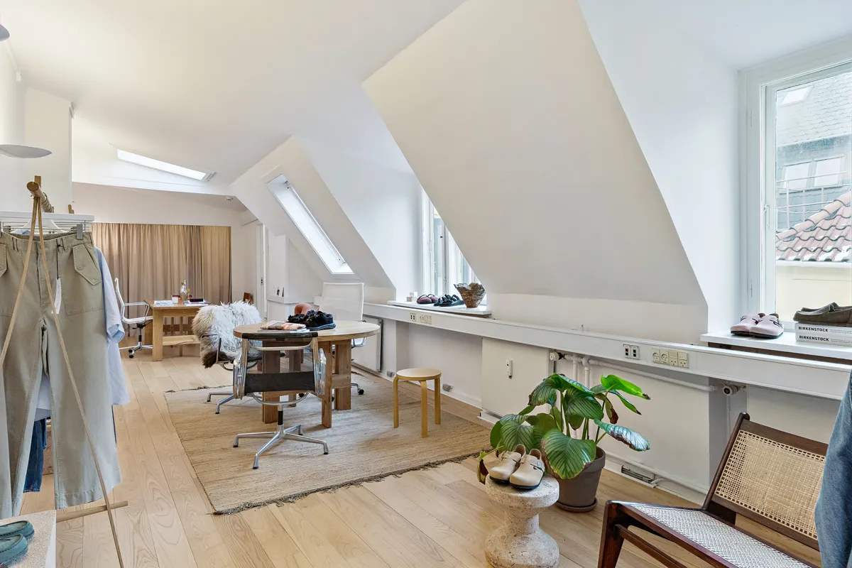 Lys kontorrum med lyse lofter, møbler og tøj på hænge og gulv samt en stor plante og sko på en fodskammel