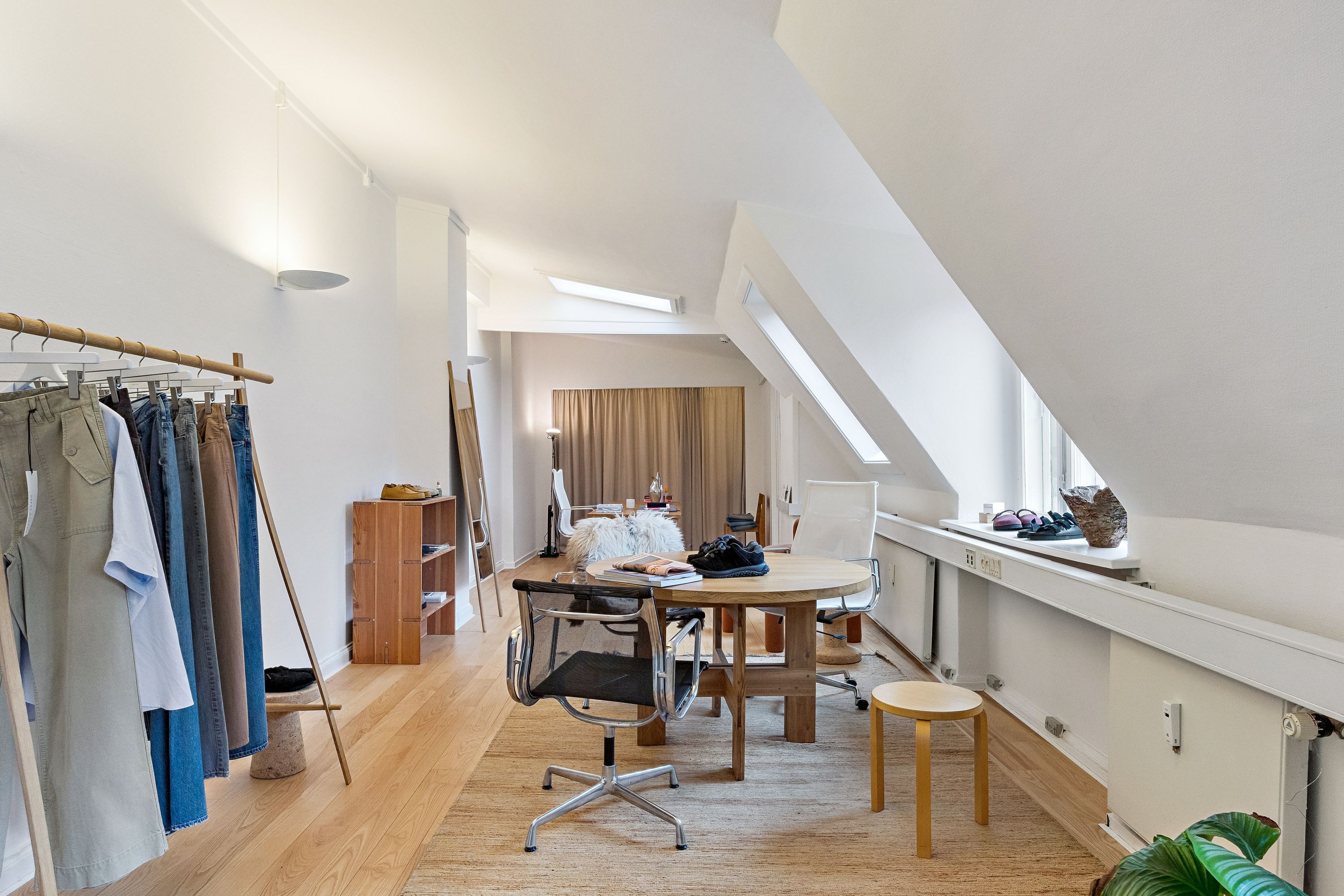 Kontor med lyse lofter og bøjet gulv med en rund træbord og to stole samt tøjstativ med tøj og sko på hylden.