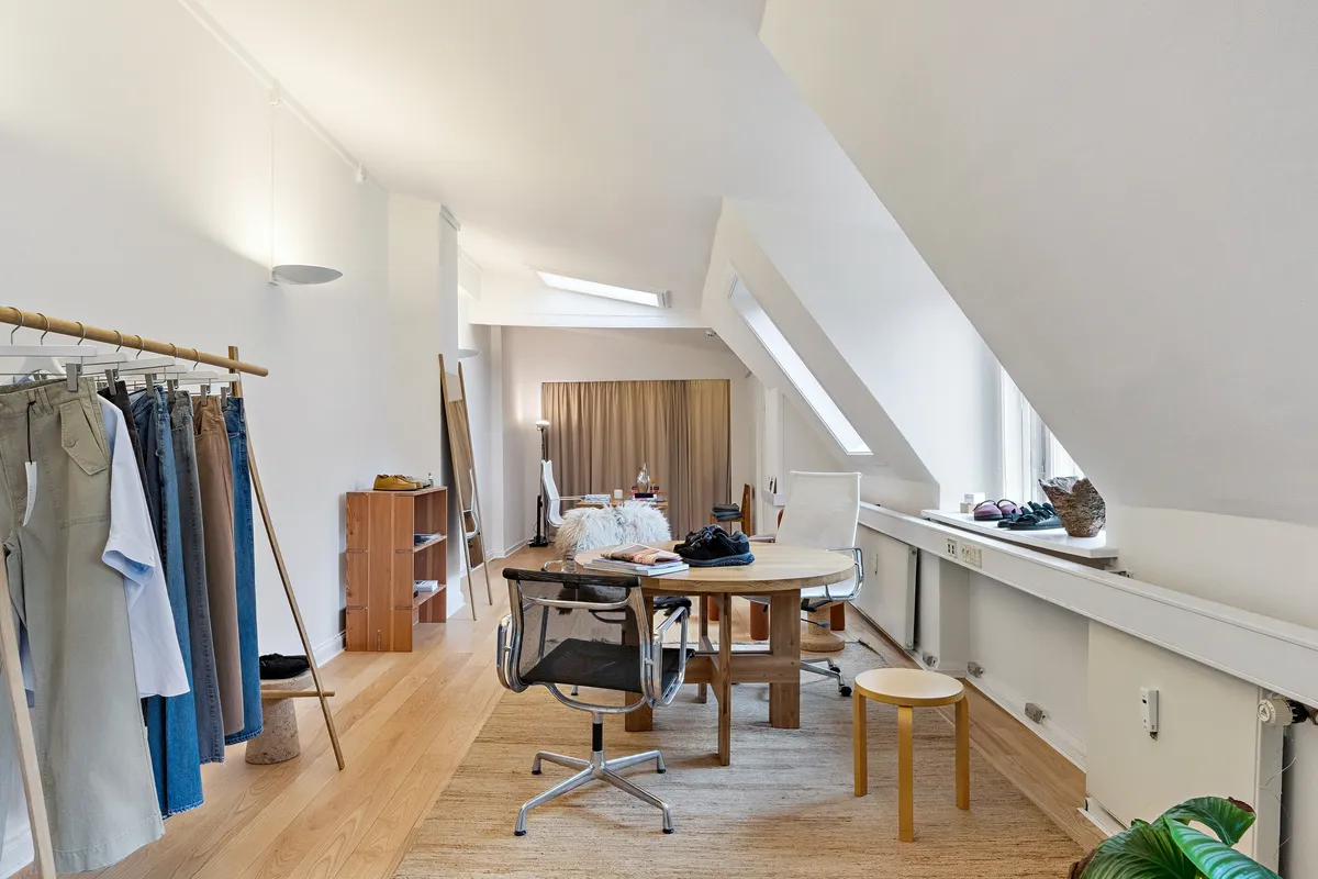 Kontor med lyse lofter og bøjet gulv med en rund træbord og to stole samt tøjstativ med tøj og sko på hylden.