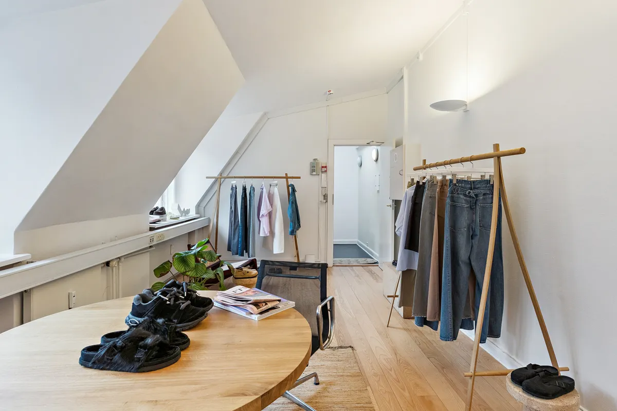 Kontor med møbler og tøj i loftsrum. Rundt bord med sko og magasiner og stole. Jeans og skjorter hængende på et træ stativ. Lys pære og plante i hjørne.