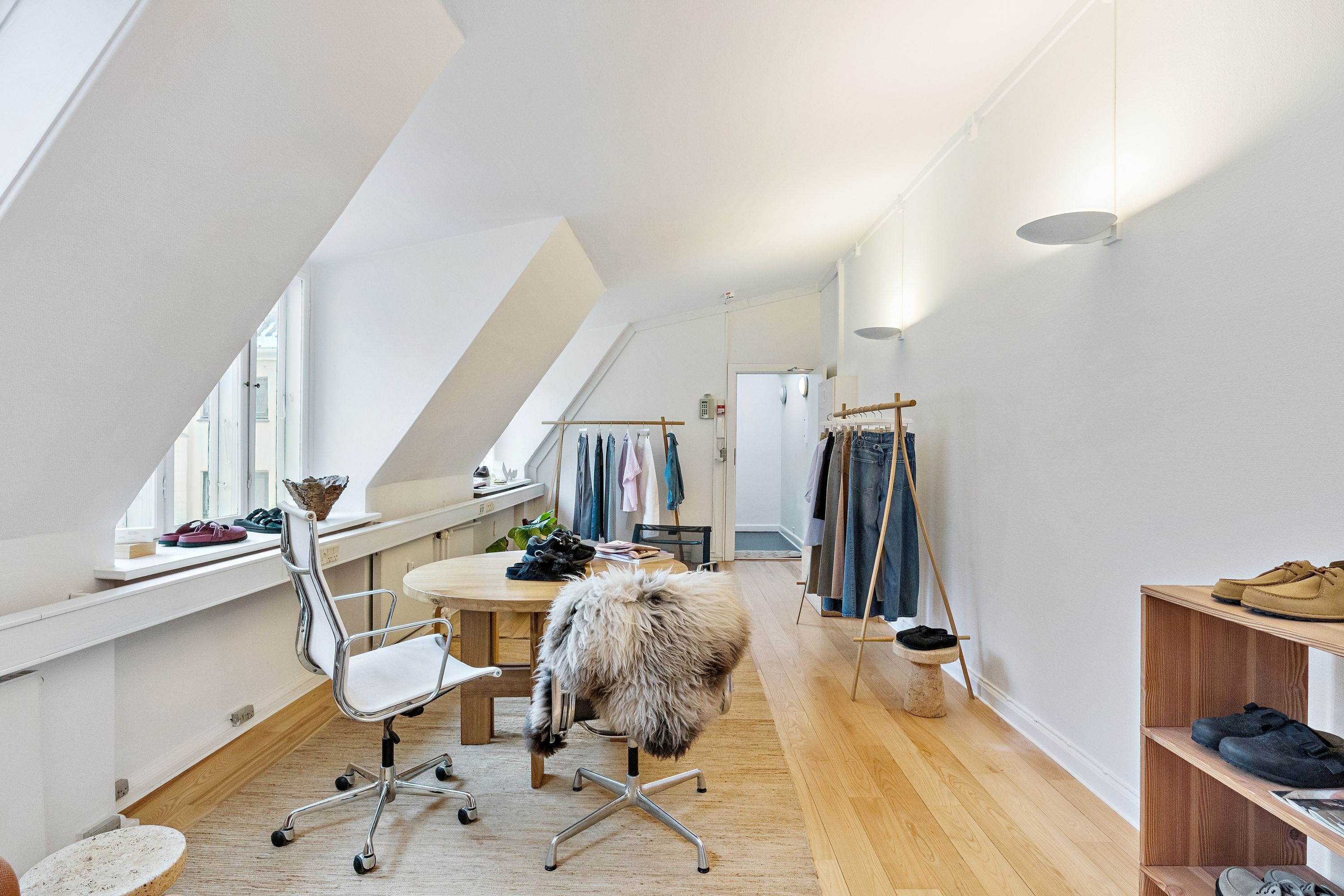 Kontor med højt til loftet og lyse loftsvinduer. Kontorbord, stol og skraldkurv. Garderobe og hængeklæder. Trægulv og træhylde med sko. Lamper og udstyr i loftet.