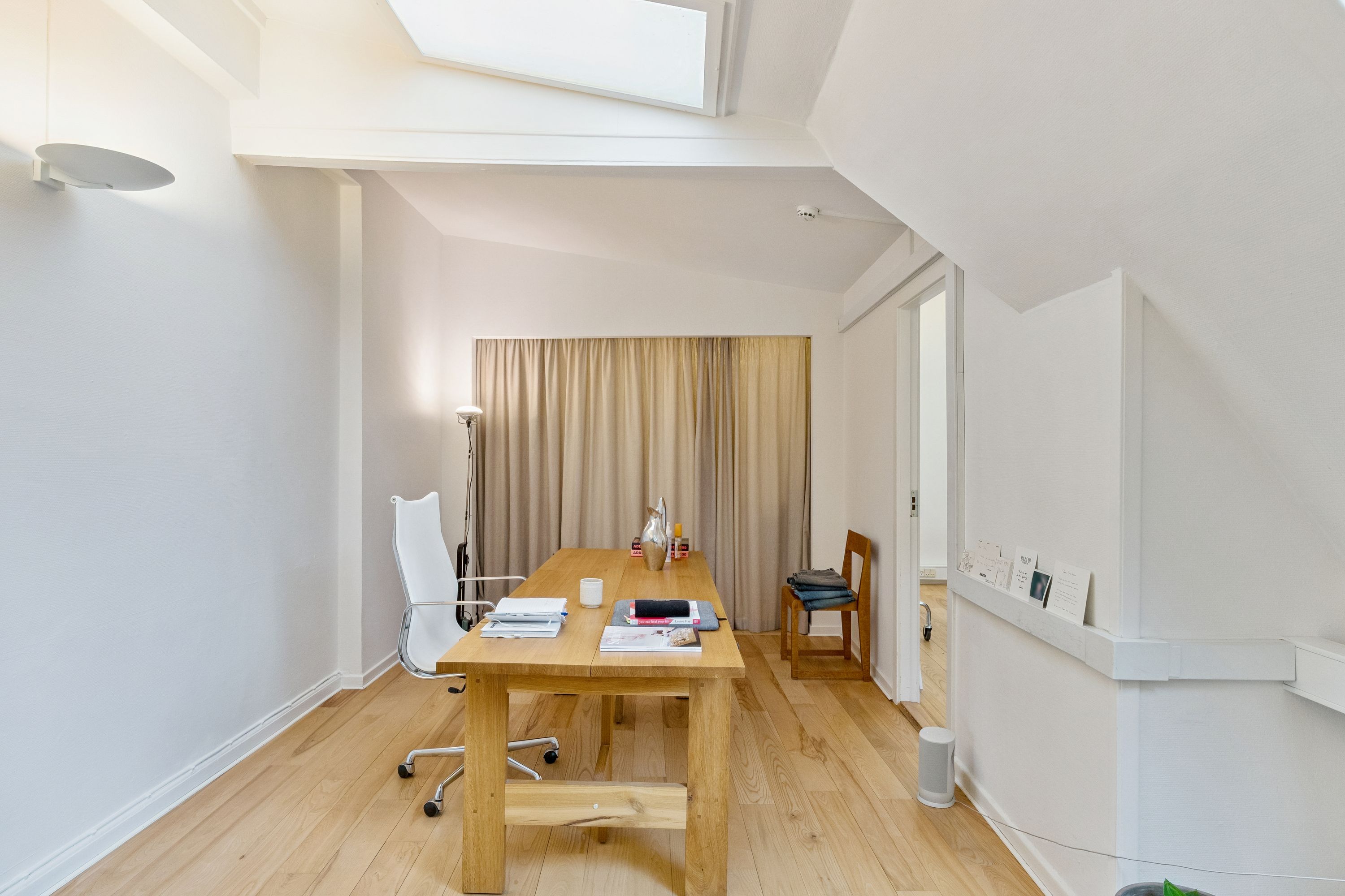 Lyst og funktionelt kontor med højloft, lysegul gardin og skrivebord med papir, kop og vase. Moderne hvidt stol med hjul og åbent garderobeskab med hængende tøj.