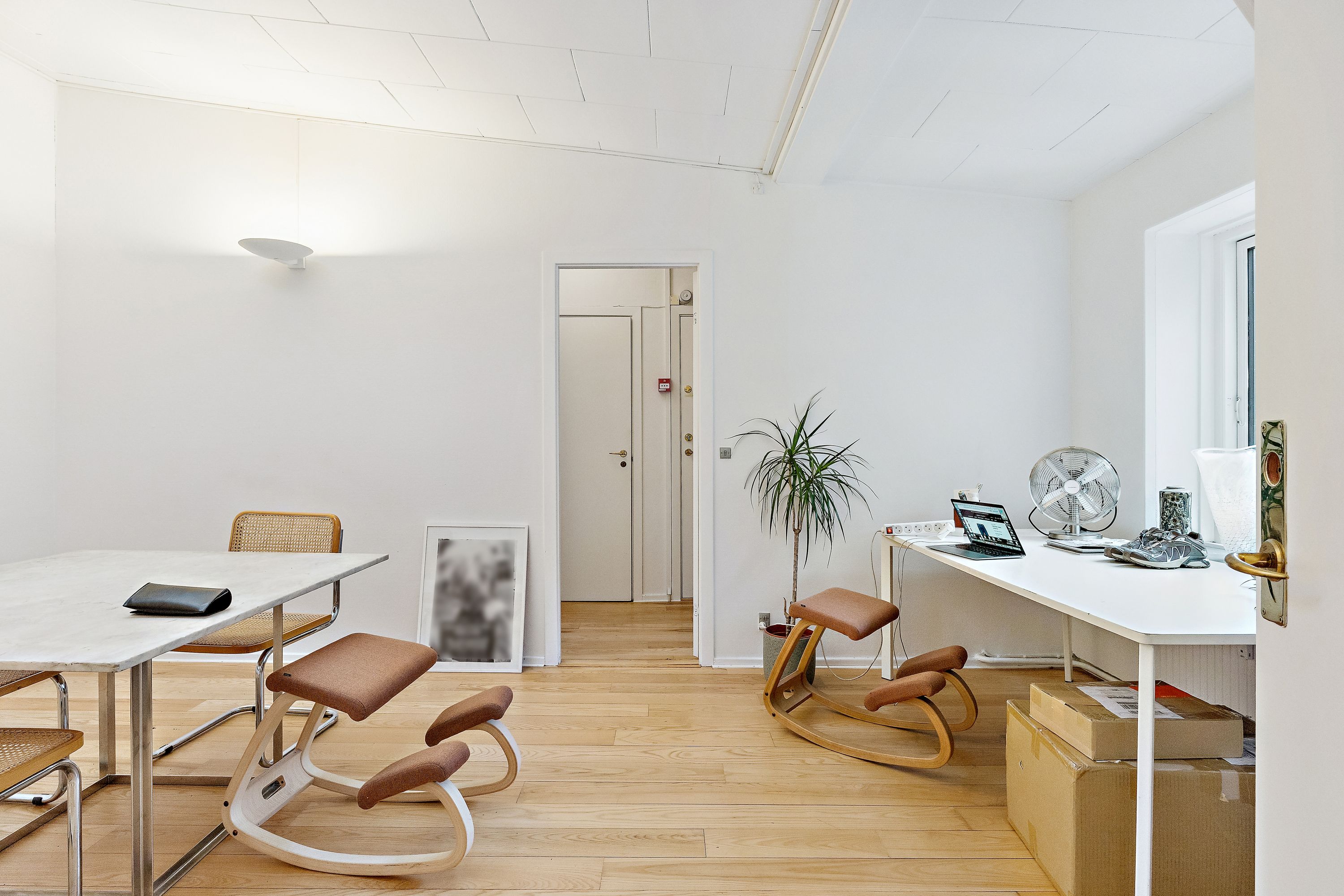 Kontor med hvide vægge og trægulve med bord og stole. Et højt skrivebord med computer, sæde og ventil. En plante og flere kasser er placeret på siden af skrivebordet. Et lysekron er hængende på loftet.