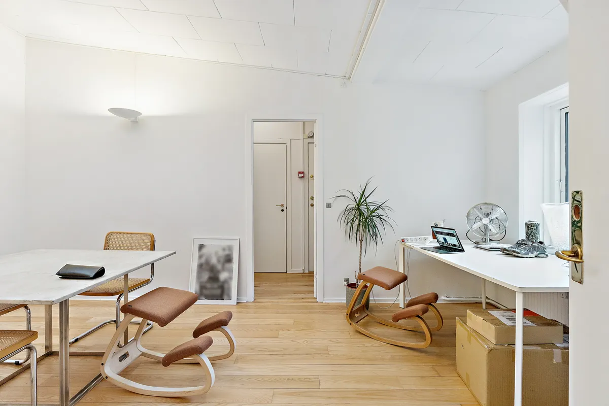 Kontor med hvide vægge og trægulve med bord og stole. Et højt skrivebord med computer, sæde og ventil. En plante og flere kasser er placeret på siden af skrivebordet. Et lysekron er hængende på loftet.