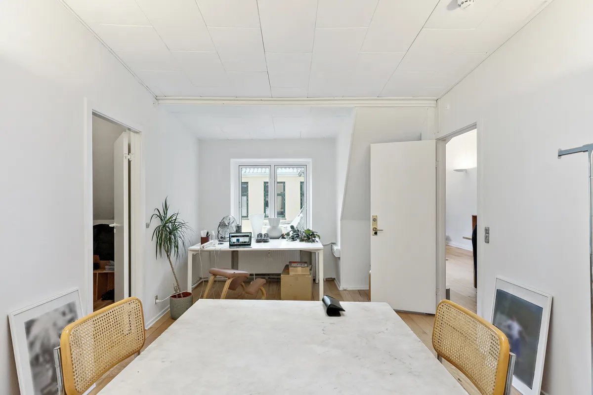 Kontor med højloft og hvidt interiør, skrivebord med bærbar computer og plante, hængelampe og højstole ved langt bord. Lyse trægulve og lukket hvidt dørblad til andet rum.
