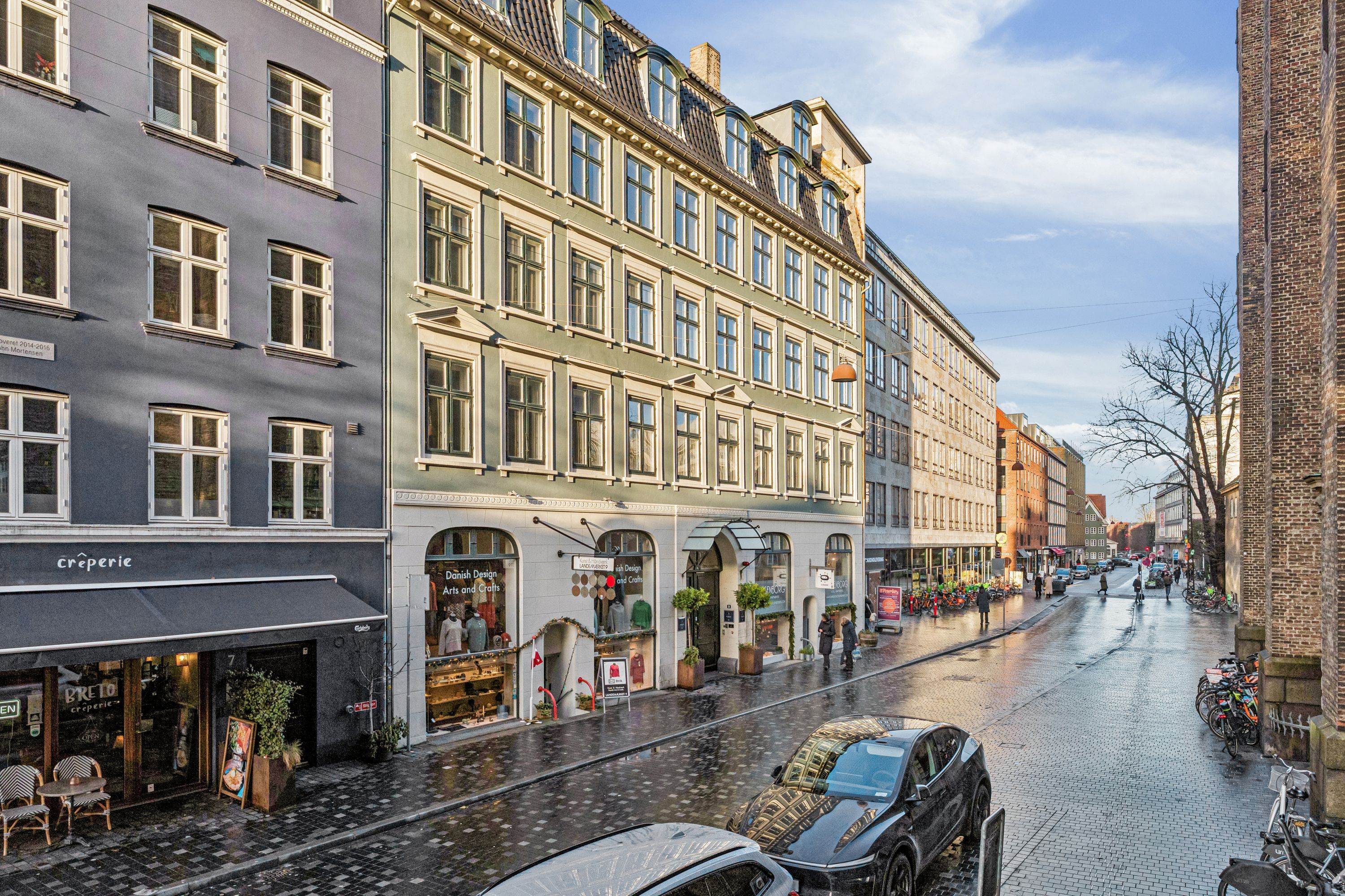 Vej med biler, cykler og mennesker, udendørs butikker med udsalg, en regnfuld dag og en bygning med mange vinduer