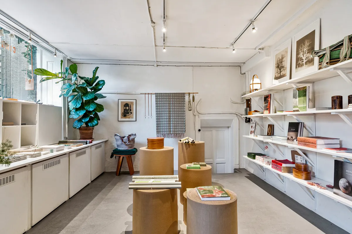 Butik med lyse loftslampe og hvidt vægmaleri. Køkken med fyldte hylder og grønne planter. Fikse træhylder med bøger og kunstværker. Korte cylinderhylder med papir og stoffer.