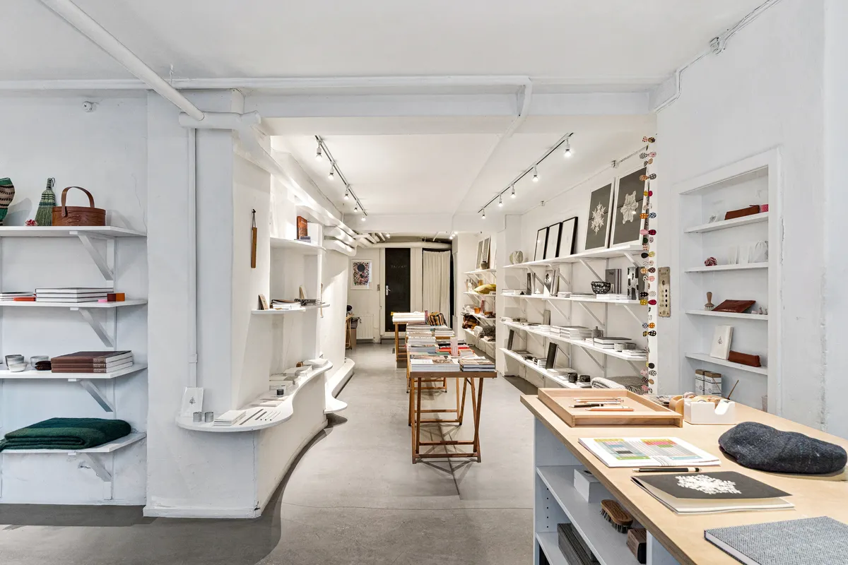 Minimalistisk butiksinteriør med hvide vægge, høje hylder, lysekroner og flere skriveborde med papirvarer og bøger. Alle hylder er fyldt med bøger, og der er en lyseblå kuffert på gulvet