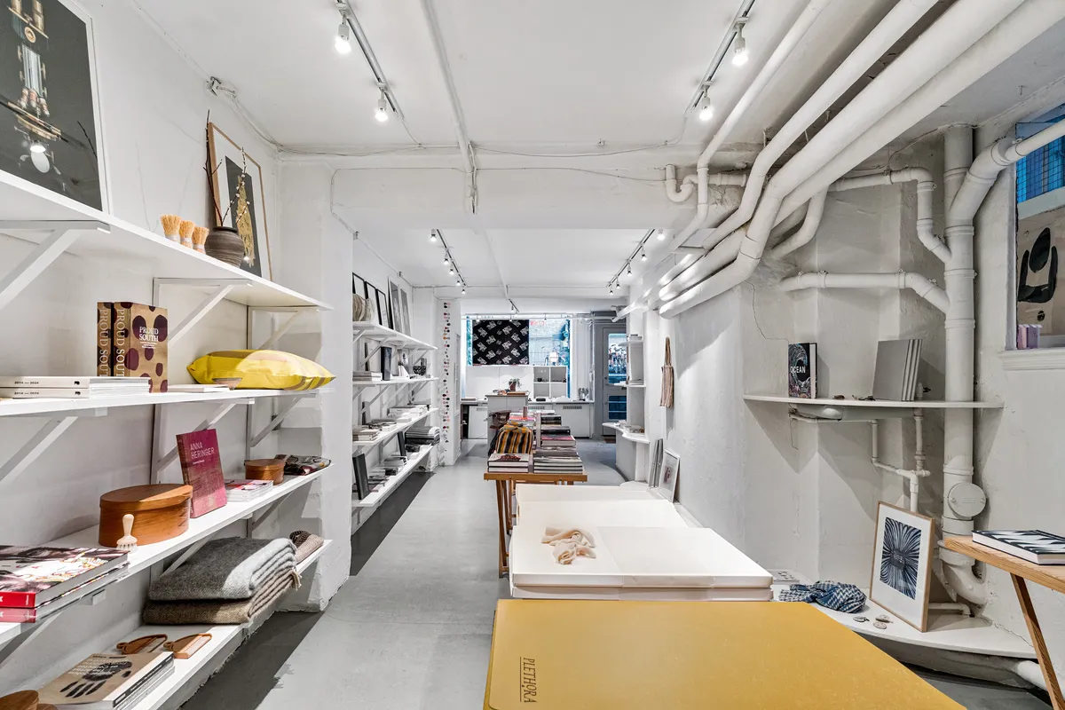 Butikslokaler med hylder og møbler, indretning med kunstværker og bøger, minimalistisk design med hvide vægge, åbent rum med lysekroner og røre, indretning med tekstiler og trægenstande.