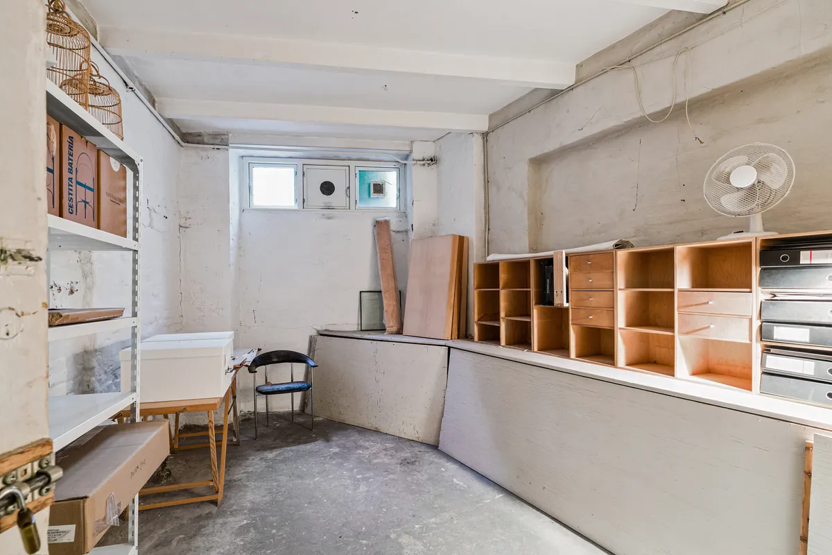 Lagerrum til leje med høje hylder, skrivebord, stol og kasser samt fan, der står på loftet i København K