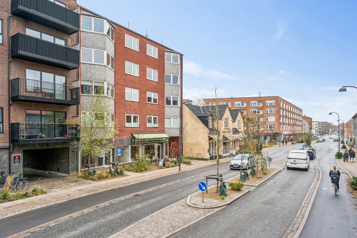 Mennesker går på fortovet i en gågade. En kvinde kører på cykel. Der er flere biler, der kører langs vejen. Bygninger med butikker og parkerede biler. Træer og buske langs vejen.