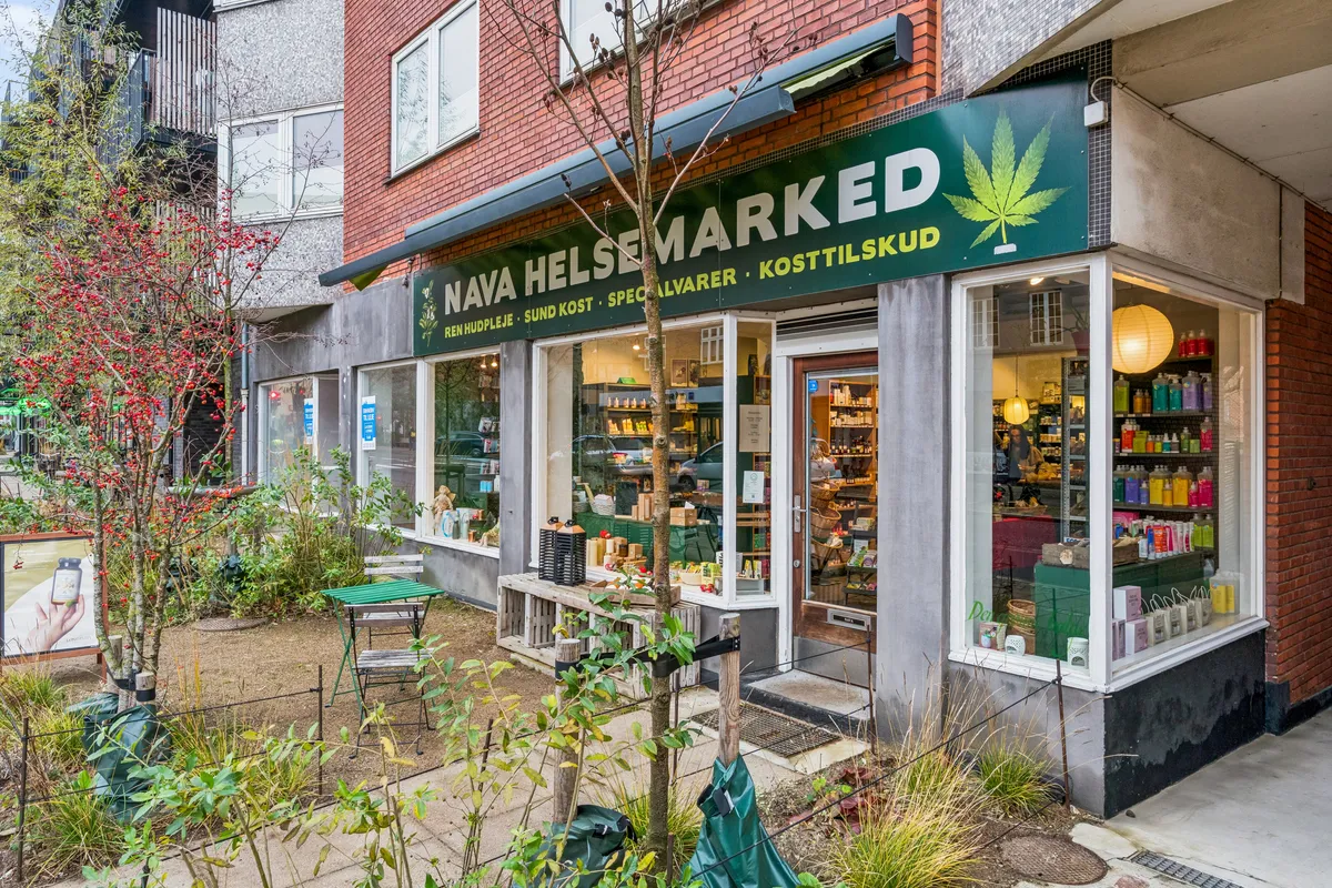 Butik med grønt skilt med teksten Nava Helsmarked og et grønt marijuanablad samt produkter i vinduet, udendørsborde og en grøn plante