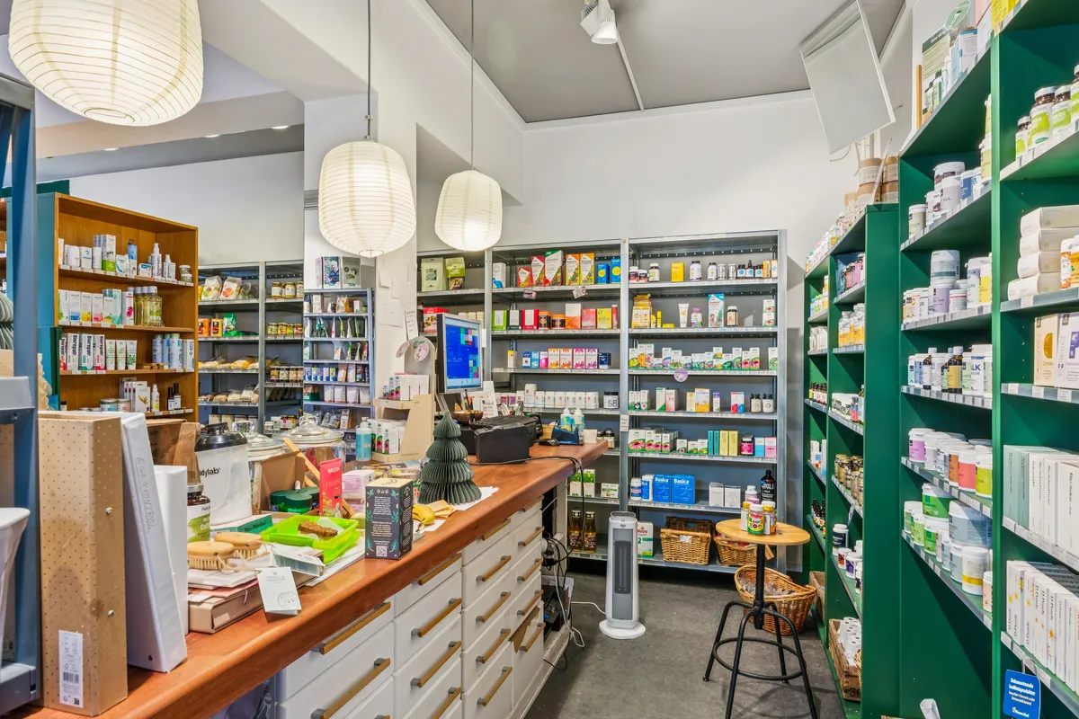 Lyst butikslokale med solskinsgule lanterner og lyse stolpehyggelamper, hylder med kosmetik og sundhedsprodukter, modtagerpult med computer og stol, køkken med højdisk og hjemmelavet juledekor