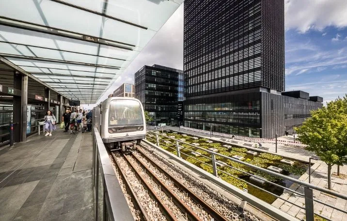 Moderne metrostation med tog, glastag og høje bygninger bagved, folk med cykler og en grøn plante på perronen