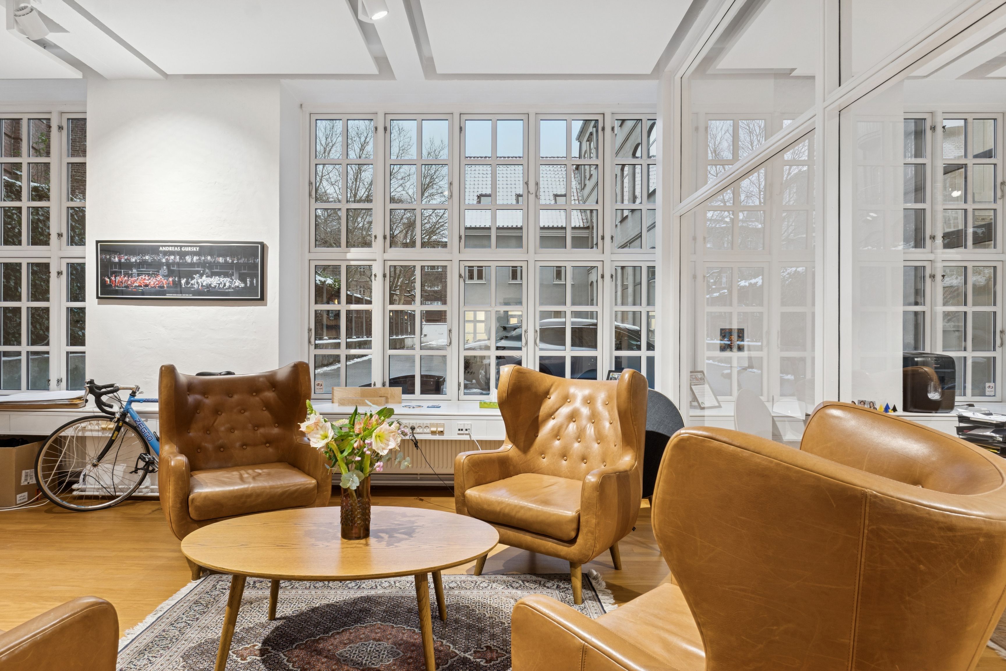 En lys og moderne kontorlounge med læderstole, et rundt bord, en blomstervase og en cykel i baggrunden. Store vinduer giver masser af naturligt lys og udsigt til gaden.